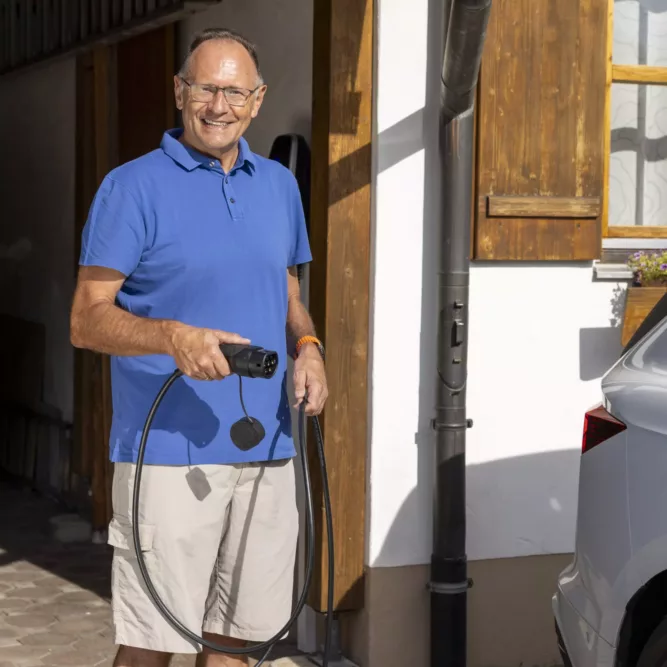 Haus der Energie – Elektroauto zu Hause mit Photovoltaik laden Mann lädt sein Elektroauto mit einem Ladekabel vor seinem Haus mit Photovoltaik von Haus der Energie